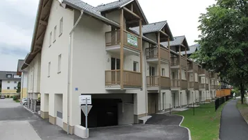 Modernes Mehrfamilienhaus mit hellen Fassaden, zahlreichen Balkonen und Tiefgaragenzufahrt, umgeben von Grünflächen und Bäumen.