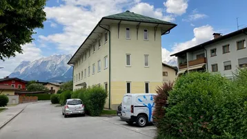 Gelbes Mehrfamilienhaus mit grünem Dach und Bergblick in ländlicher Umgebung.