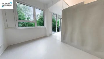 Helles Zimmer mit großen Fenstern, Blick ins Grüne und Zugang zu einem Balkon.