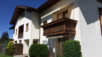 Gepflegtes Mehrfamilienhaus mit weißer Fassade, Holzbalkonen und grüner Umgebung unter blauem Himmel.