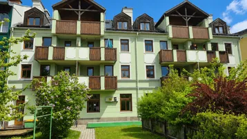 Mehrfamilienhaus mit hellgrüner Fassade, Holzbalkonen und Dachgauben unter blauem Himmel.