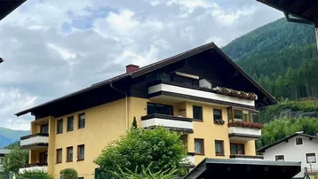 Mehrfamilienhaus in alpiner Umgebung mit Balkonen und Satteldach, umgeben von grünen Bäumen.