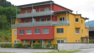 Modernes Mehrfamilienhaus mit leuchtend rot-gelber Fassade und mehreren Balkonen in grüner Umgebung.