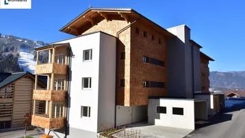 Modernes Apartmentgebäude mit Holzschindelfassade und Balkonen in alpiner Umgebung mit Bergblick.