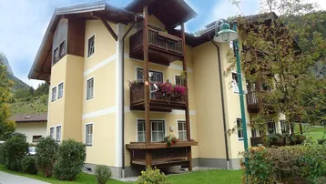 Mehrfamilienhaus mit gelber Fassade, Holzbalkonen und grüner Umgebung in den Bergen.