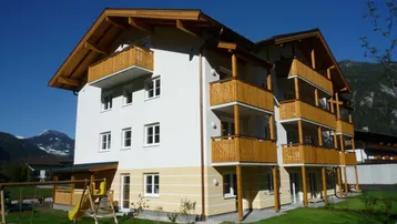 Modernes Mehrfamilienhaus mit Holzbalconen, einem kleinen Spielplatz und Bergblick im Hintergrund.
