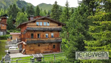 Traditionelles Holzhaus in alpiner Umgebung, umgeben von grünen Bäumen und Bergen.