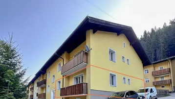 Gelbes Mehrfamilienhaus mit Holzbalkonen und parkenden Autos davor, umgeben von Bäumen.