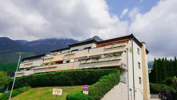 Mehrfamilienhaus mit Balkonen und Bergblick, umgeben von grünen Hecken.