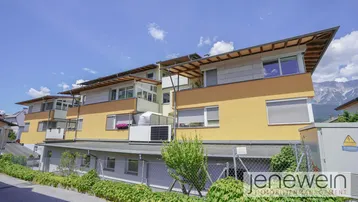 Mehrfamilienhaus mit gelber Fassade, Balkonen und Blick auf die Berge.
