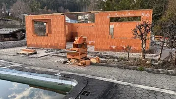 Rohbau eines Hauses mit Ziegelwänden und sichtbaren Fensteröffnungen, daneben ein Pool mit Wasserspiegelung.