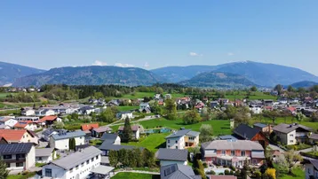 Panoramablick über eine Wohnsiedlung mit grünen Feldern und Bergen im Hintergrund unter blauem Himmel.