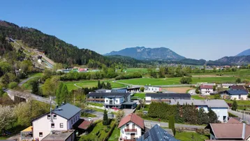 Luftaufnahme einer Wohnsiedlung mit grüner Landschaft und Bergen im Hintergrund unter blauem Himmel.