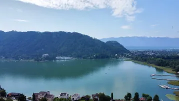 Panoramablick auf den See mit umliegenden Bergen und Häusern am Ufer unter blauem Himmel.