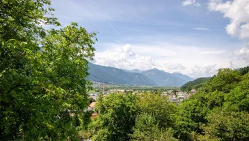 Weitläufiger Blick über ein grünes Tal mit Bergen und einer kleinen Ortschaft unter blauem Himmel.