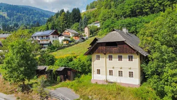 Traditionelles Haus in alpiner Umgebung mit grünen Hügeln und bewaldeten Bergen im Hintergrund.