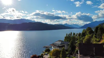 Panoramablick auf den See und die umliegenden Berge mit mehreren Häusern am Ufer an einem sonnigen Tag.