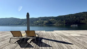 Große Holzterrasse am See mit Liegestühlen und Bergblick unter blauem Himmel.