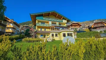 Traditionelles Gebäude in den Bergen mit gelber Fassade und grüner Dachgestaltung, umgeben von gepflegtem Garten.