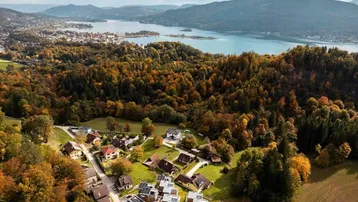 Luftaufnahme eines Dorfes mit See und Bergen im Hintergrund, umgeben von herbstlichen Wäldern.