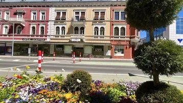 Bunte Blumenbeete im Vordergrund mit historischen Gebäuden an einer belebten Straße.