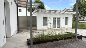Moderne Holzterrasse mit Glasüberdachung und Blick auf ein weißes Nebengebäude.