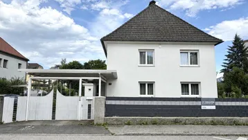 Weißes Einfamilienhaus mit Satteldach, Carport und Vorgarten, bereit zum Verkauf.