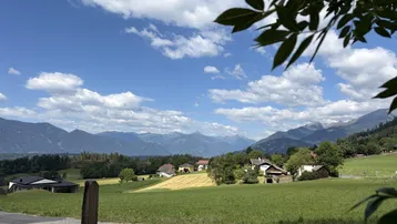 Malerischer Blick auf eine ländliche Landschaft mit Bergen, grünen Feldern und vereinzelten Häusern unter blauem Himmel.