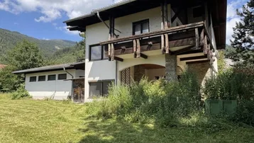 Traditionelles Haus mit Balkon, umgeben von üppiger Vegetation und Bergen.