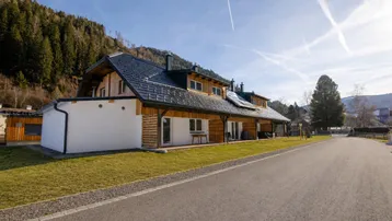 Modernes Gebäude mit Holzfassade und dunklem Dach, umgeben von Grünflächen und Bergen unter blauem Himmel.