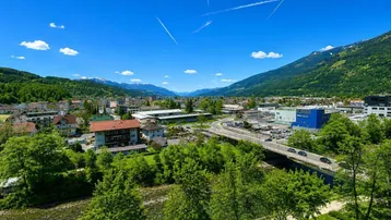 Weitläufiger Blick über eine Stadt mit Fluss, grünen Bäumen und schneebedeckten Bergen im Hintergrund.