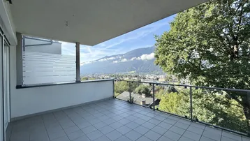 Großer Balkon mit Fliesenboden, Glasgeländer und weitem Blick auf Berge und Stadt.