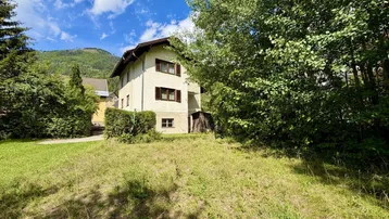 Freistehendes Haus mit heller Fassade, umgeben von viel Grün und Bäumen in bergiger Landschaft.