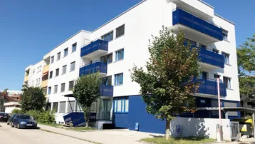 Modernes Mehrfamilienhaus mit weißer Fassade und blauen Balkonen unter blauem Himmel.