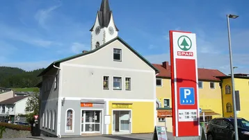 Gebäude mit gemischter Nutzung, einem SPAR-Schild und einem Kirchturm im Hintergrund unter blauem Himmel.