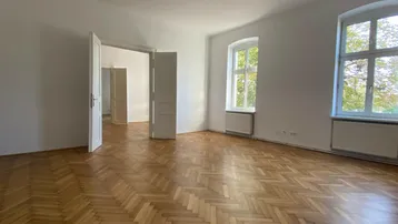 Geräumiges Wohnzimmer mit Fischgrätparkett, hohen Decken und großen Fenstern, die viel Tageslicht hereinlassen.