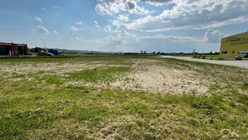 Weitläufiges, unbebautes Grundstück mit Gras und Erde, umgeben von Gewerbegebäuden unter einem bewölkten Himmel.