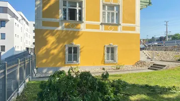 Gelbes Gebäude mit traditioneller Architektur, grünen Balkonen und einem gepflegten Rasenbereich im Vordergrund.
