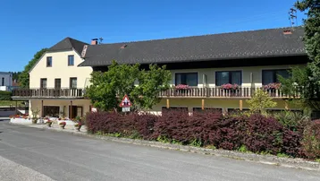 Großes Gebäude mit mehreren Balkonen, umgeben von grünen Büschen und einer Straße.
