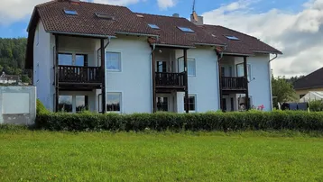 Mehrfamilienhaus mit weißer Fassade, mehreren Balkonen und Satteldach, umgeben von Grünflächen und Hügeln.