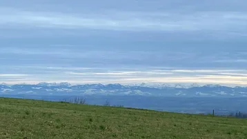 Weitläufiger Blick über grüne Hügel auf eine beeindruckende Bergkette am Horizont unter bewölktem Himmel.