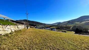 Weitläufige grüne Landschaft mit sanften Hügeln und vereinzelten Häusern unter blauem Himmel.