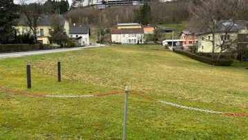 Grünfläche mit umliegenden Häusern und bewaldetem Hügel im Hintergrund unter bewölktem Himmel.