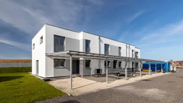 Moderne Reihenhäuser mit weiß-grauer Fassade, Carport und Grünfläche unter blauem Himmel.