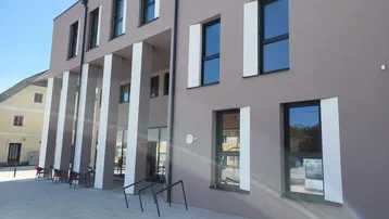 Moderne Gebäudefassade mit großen Fenstern, Säulen und einem gepflasterten Vorplatz bei sonnigem Wetter.