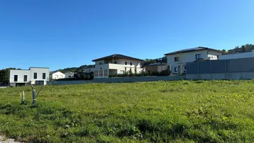Grünes Baugrundstück mit Blick auf moderne Wohnhäuser und eine hügelige Landschaft unter blauem Himmel.
