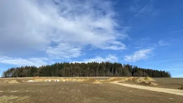 Weitläufiges Grundstück mit Erdhügeln und angrenzendem Wald unter blauem Himmel.