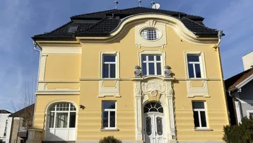 Historisches Gebäude mit gelber Fassade, reich verzierten Details und Mansarddach unter blauem Himmel.