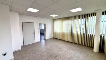 Geräumiges Büro mit hellem Linoleumboden, abgehängter Decke und vertikalen Jalousien an den Fenstern.