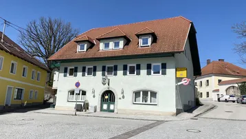 Traditionelles Gebäude mit hellgrüner Fassade, roten Dachziegeln und gepflastertem Vorplatz in sonniger Lage.